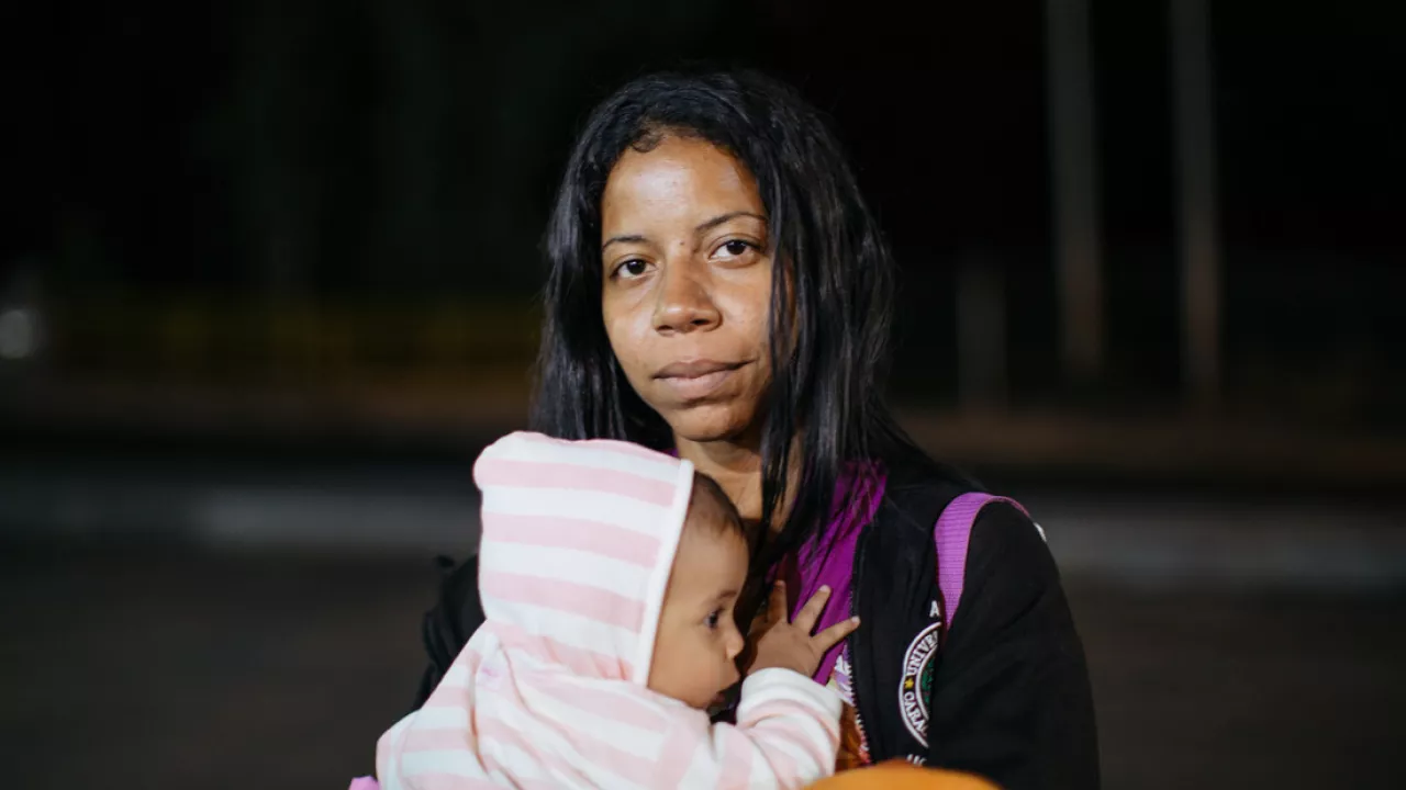 A Venezuelan woman looks at the camera while holding a small child in her arms. The baby is wearing a light pink and white striped hoodie onesie.